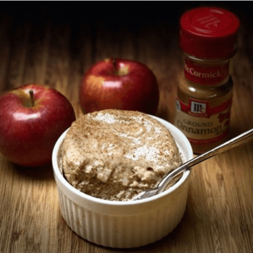 Eine Schale Apfel-Tassenkuchen mit Protein neben zwei Äpfeln und einem Glas Zimt
